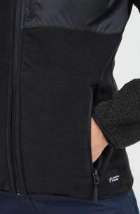 Veste polaire noire avec un panneau supérieur lisse. Comprend une poche zippée et une étiquette de logo. Tissu texturé sur tout le corps. Matériel chaud.