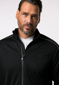 Chaqueta negra con cremallera y textura suave, que cuenta con un cuello alto, camiseta blanca debajo y un sutil detalle del logo en el frente.