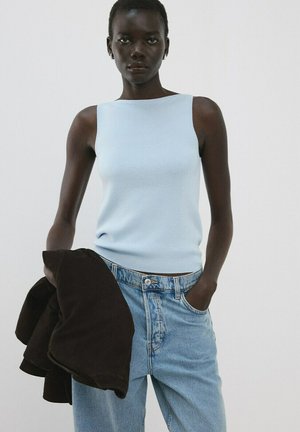 Frau trägt ärmelloses hellblaues Oberteil und hoch taillierte hellblaue Jeans, hält eine schwarze Jacke in einer Hand vor weißem Hintergrund.