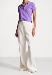 Kvinna iklädd en kortärmad lila pikétröja instoppad i högmidjade vida vita byxor med krämfärgade skor, stående mot en vit bakgrund.