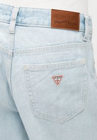 Πίσω τσέπη από ανοιχτό μπλε denim με τρίγωνο λογότυπο Guess και καφέ δερμάτινη ετικέτα μάρκας στη μέση.