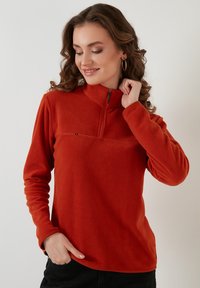 Fleece pulóver s čtvrtovým zipsom v živých červeno-oranžových farbách, s textúrovanou panelou cez hrudník a priliehavým strihom s dlhými rukávmi.