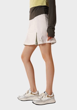 Persona che indossa una gonna plissettata bianca, una maglia a maniche lunghe a righe e sneaker bianche con dettagli beige e argento, in piedi contro uno sfondo semplice.