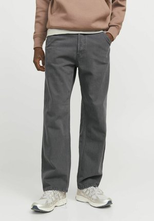 Homme portant un pantalon droit gris, un sweat à capuche beige et des baskets blanches, debout avec une main dans la poche devant un fond uni.