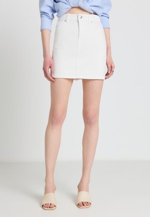 Femme portant une mini-jupe blanche, une chemise bleue claire à manches longues nouée et des sandales beiges à bout ouvert, debout devant un fond uni.
