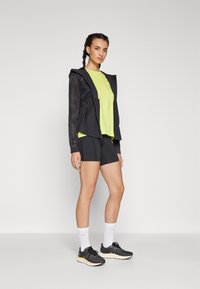 Frau, die in schwarzen Sportshorts, einem limonengrünen T-Shirt, einer schwarzen Zip-Jacke, weißen Socken und schwarzen Laufschuhen vor einem schlichten Hintergrund steht.