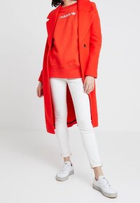 Abrigo largo rojo sobre un sudadera roja con logotipo blanco, combinado con jeans blancos y zapatillas grises, con un diseño limpio y minimalista.