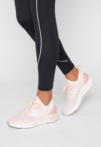 Lichte sneakers in roze en wit, met een textuur bovenkant, elastische veters en een gevoerde zool. Gecombineerd met zwarte legging.