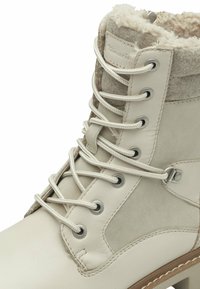 Bottine beige avec une tige en cuir lisse et en feutre doux, ornée de lacets blancs, d'œillets en métal et d'une semelle en caoutchouc. Détail de col texturé.