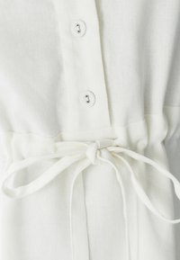 Blusa blanca de tela ligera con botones en la parte delantera, cintura con cordón y un acabado texturizado.