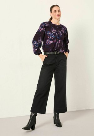 Schwarze, hoch taillierte Wide-Leg-Jeans kombiniert mit einer lila blumigen Bluse mit langen Ärmeln und Knöpfen. Das Model trägt schwarze, knöchelhohe Stiefel mit Absatz.