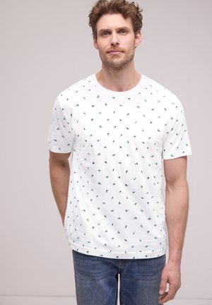 Homme aux cheveux bouclés portant un t-shirt blanc à manches courtes avec un motif de petites feuilles vertes et noires, et un jean bleu, debout devant un fond uni.