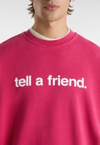 Homme portant un sweat-shirt rose vif avec le texte blanc « tell a friend. » sur le devant, col de chemise blanche visible en dessous.