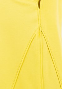 Primo piano di tessuto giallo con pieghe triangolari cucite simmetricamente che creano un dettaglio strutturato sul materiale.