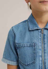 Denim srajca v svetlo modri barvi s kratkimi rokavi, sprednjim zadrgo in žepom na prsih. Ima klasičen ovratnik in zbledel videz tkanine.