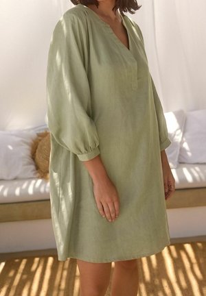 Žena stojící uvnitř, oblečená v volných, světle zelených šatech z lnu s dlouhým rukávem a výstřihem do V, se zapnutými rukávy.