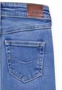 Blå denimjeans med en brun läderetikett på midjan. Har en enda bakficka med ett distinkt, böjt sy mönster.