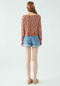 Strickpullover mit Rautenmuster in Orange und Weiß, kombiniert mit Jeansshorts. Das Modell trägt hellbraune, hölzerne Clogs mit Absatz und ein passendes Stirnband.