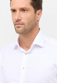 Man met lichtbruine stoppels in een wit overhemd, die iets naar rechts kijkt tegen een effen witte achtergrond.