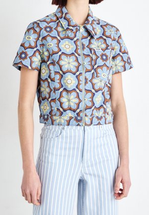 Žena nosí krátkou bundu na zip s krátkými rukávy s květinovým vzorem v modré, hnědé a žluté barvě, kombinovanou s světle modrými a bílými pruhovanými kalhotami.