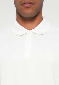 Camisa polo branca com colarinho clássico, pala de três botões e um logotipo discreto no peito, com uma textura de algodão suave.