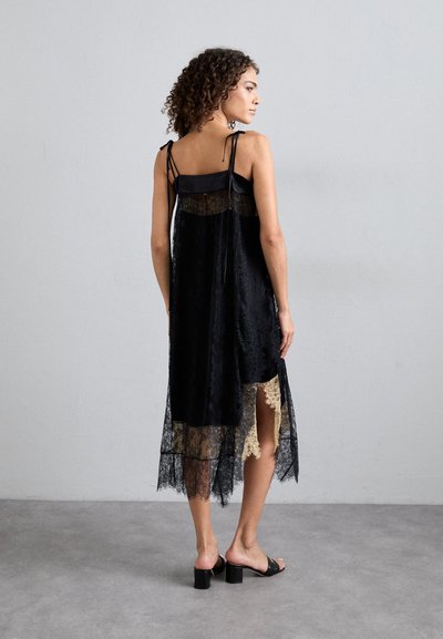 Μαύρο μίντι φόρεμα από δαντέλα με διαφανή υφή, που διαθέτει μπεζ δαντελένια περίγραμμα, ρυθμιζόμενα ραμμένα λουριά και πλαϊνό σκίσιμο για επιπλέον λεπτομέρεια.