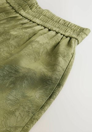 Lichtgroene stof met een textuur van een bloemenpatroon, voorzien van een geplooide elastische tailleband en een gladde afwerking. Close-up zicht benadrukt de details.