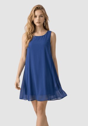 Joven mujer con cabello largo y rubio, vestida con un vestido azul sin mangas en forma de A, de pie contra un fondo liso gris claro.