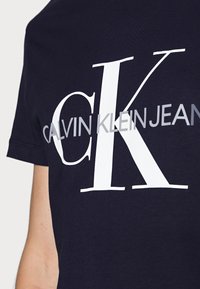 Námořnická modrá bavlněná tričko s velkým bílým grafickým nápisem "CK" a "CALVIN KLEIN JEANS" v šedé barvě, krátké rukávy, kulatý výstřih.