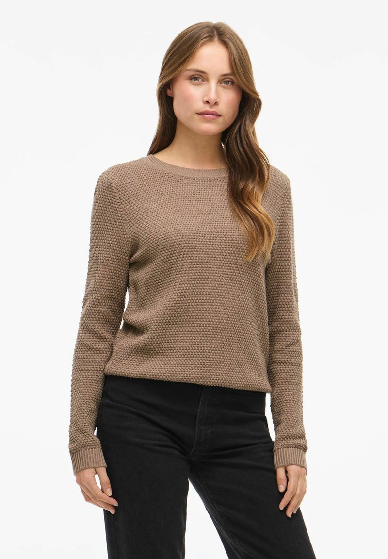 Maglione marrone chiaro textured con collo a giro e maniche lunghe, abbinato a pantaloni neri. Il tessuto presenta un motivo intrecciato distintivo.