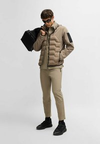 Beige vadderad jacka med huva, tillsammans med en beige topp och slim-fit byxor. Mörka sneakers och en svart väska kompletterar outfiten.