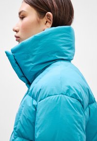 Padded jakna u svijetloj tirkiznoj boji, sa visokim ovratnikom, kviltanom teksturom i glatkom, mekanom završnom obradom. Nema vidljivih dijelova ili logotipa.
