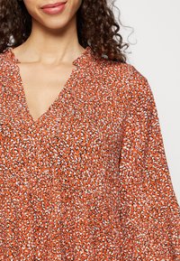Blusa laranja com padrão, decote em V. O tecido apresenta uma mistura de manchas pretas e brancas, com uma superfície ligeiramente texturada e um corte solto.