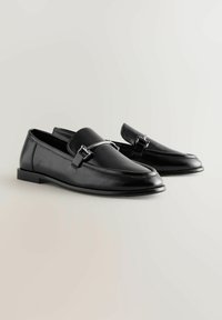 Zwarte leren loafers met een glanzende zilveren gespdetail, met een ronde neus en een lage platte hak, die een soepele textuur en een strak ontwerp tonen.