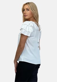 Biała bluzka z falbaniastymi rękawami, z dekoltem w kształcie litery V z tyłu i wiązanym zapięciem. Gładka tekstura i luźny krój, zestawiona z ciemnymi spodniami.