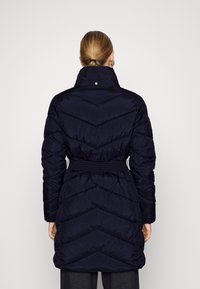 Námořnický péřový kabát s chevronovým prošíváním, páskem v pase, vysokým límcem a zapínáním na patentky. Hladká textura látky a délka do poloviny stehen.