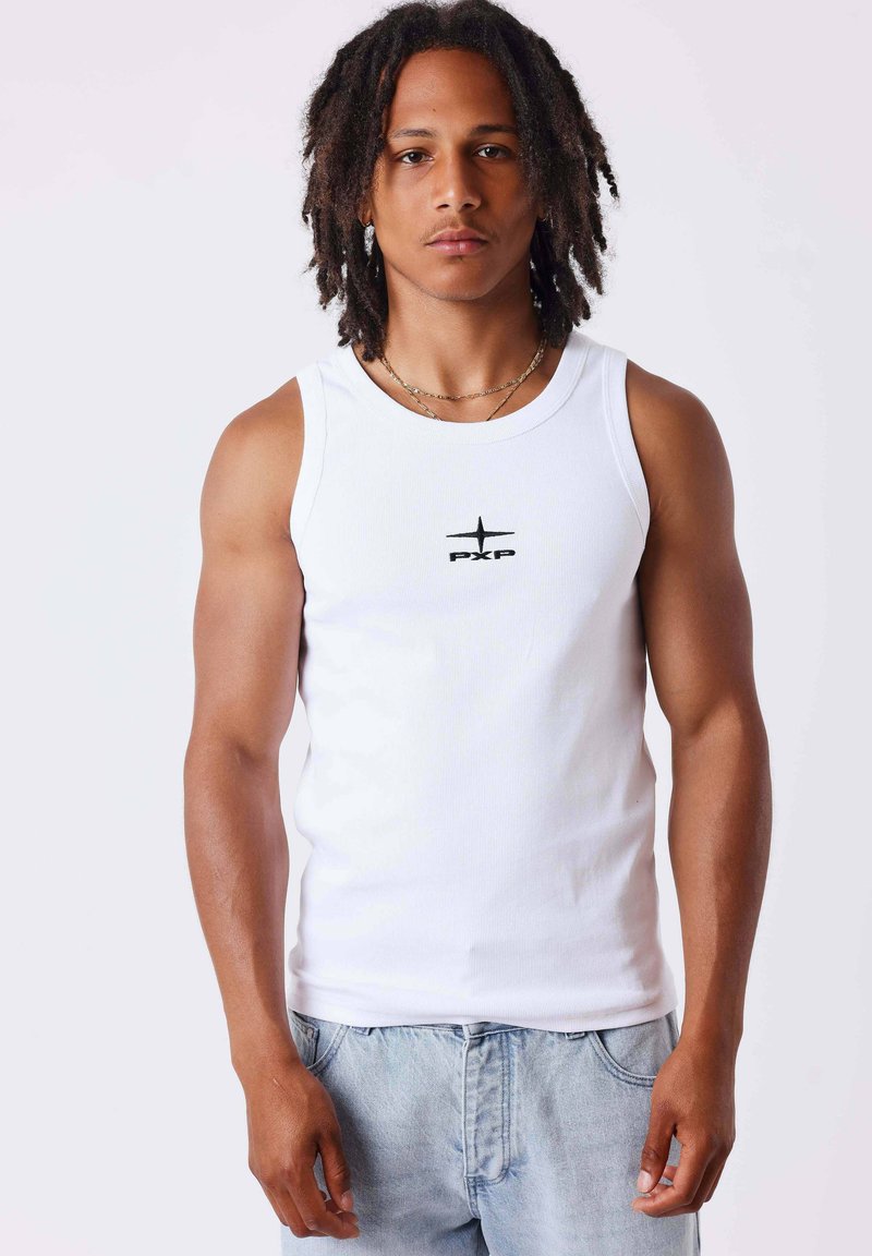 Homme avec des dreadlocks portant un débardeur blanc avec le logo "PXP" et un jean bleu clair, debout devant un fond uni.