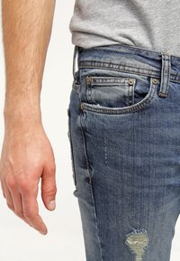 Pantalones vaqueros azules con acabado desgastado, que cuentan con un bolsillo pequeño, remaches metálicos y detalles rasgados en el muslo.