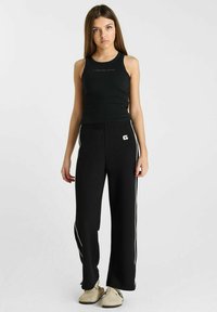 Zwarte geribbelde tanktop gecombineerd met zwarte wijde broek met witte zijstrepen en een elastische tailleband, gedragen met lichtgekleurde schoenen.