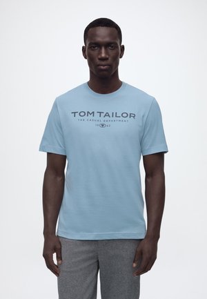 Man draagt lichtblauw Tom Tailor T-shirt en grijze broek, staand tegen een effen witte achtergrond.