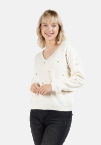 Krém színű, V nyakú pulóver, apró arany szívmintákkal, bordázott mandzsettákkal és aljjal. Sötét farmernadrág felett viselve.