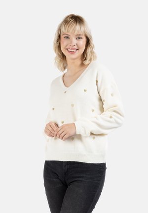 Värikäs villapaita, jossa on V-huppu, ja pienen kultaisen sydänkuvion ympäri sekä ribbireunukset hihansuissa ja helmassa. Päällä tummat farkut.