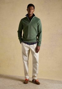 Homme debout devant un fond neutre portant un pull vert à fermeture éclair quart, une chemise à carreaux, un pantalon beige et des baskets marron avec des lacets orange.