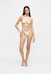 Femme debout pieds nus portant un bikini jaune avec un liseré gris sur un fond blanc uni.