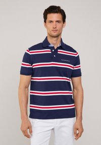 Polo azul marino con rayas horizontales rojas y blancas, de manga corta y con un cuello clásico. Presenta un pequeño bolsillo etiquetado en el pecho.