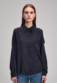 Ciemnoniebieska koszula z guzikami, wykonana z teksturowanej tkaniny, z kołnierzykiem w szpic, długimi rękawami i dekoracyjnymi guzikami na ramionach.