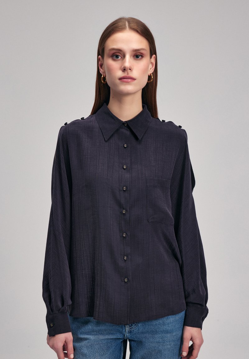 Ciemnoniebieska koszula z guzikami, wykonana z teksturowanej tkaniny, z kołnierzykiem w szpic, długimi rękawami i dekoracyjnymi guzikami na ramionach.