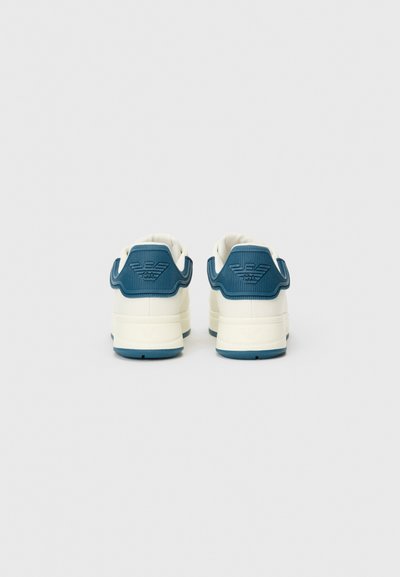 Vita sneakers med en texturerad blå accent vid hälen, med logotypdetalj och en gummisula. Bakvy som visar sömmar och designelement.