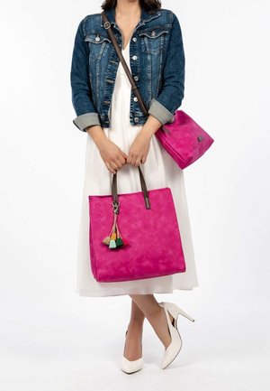 Rosa geflochtene Tragetasche mit braunen Griffen und einem bunten Quastenakzent, kombiniert mit einem passenden kleinen Beutel. Das Modell trägt eine Jeansjacke.