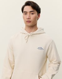 Kremowa bluza z kapturem wykonana z miękkiego materiału, z kieszenią z przodu, sznurkiem do regulacji kaptura i haftowanym niebieskim logo na lewej piersi.